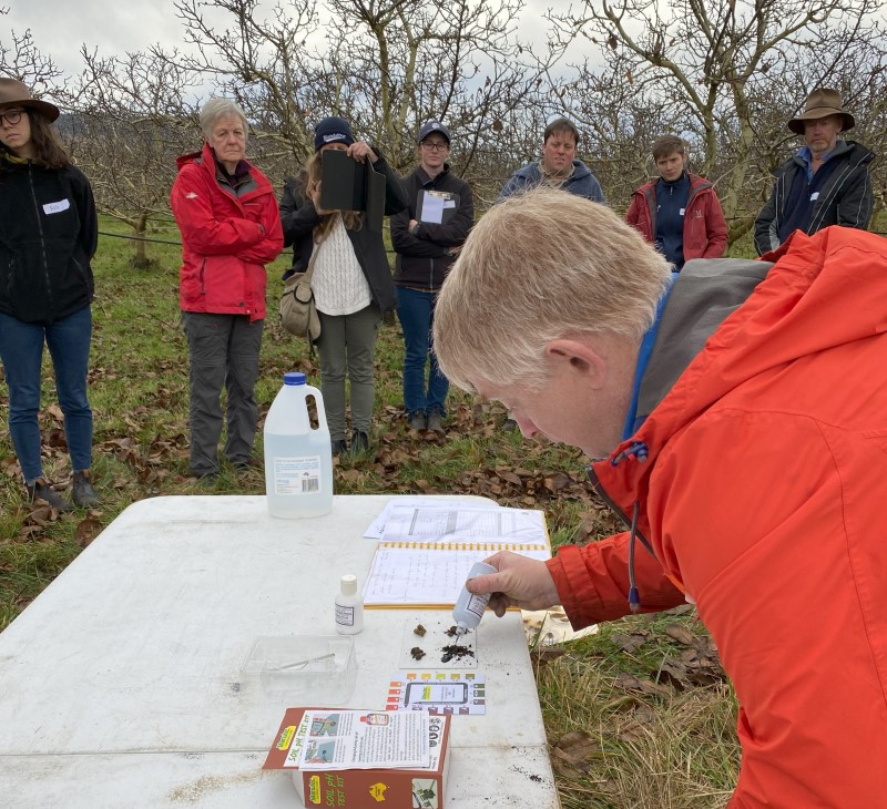 People stand in an orchard around a table with a pH test kit. A man is applying drops of solution to soil samples. 