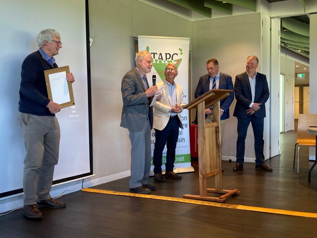 Five men stand by a lectern, one holding a microphone and one holding a framed certificate. 